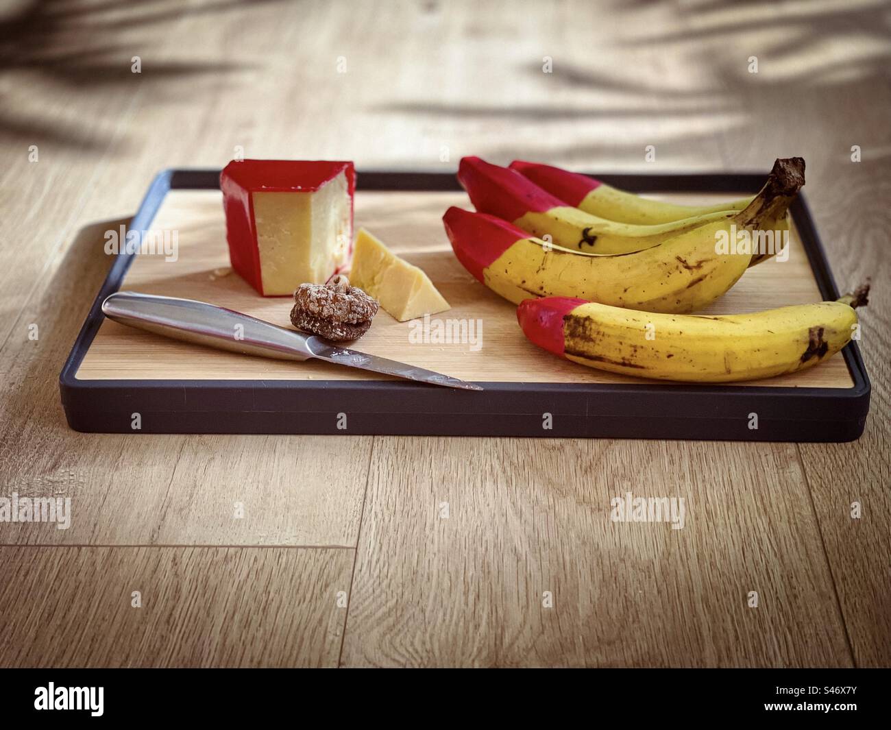 Aged cheddar cheese with red wax coating, a dried fig, organic, red wax tipped bananas and a knife on wooden cutting board on table with copy space. Sunlight and shadows. - Smartphone Captured Stock Image
