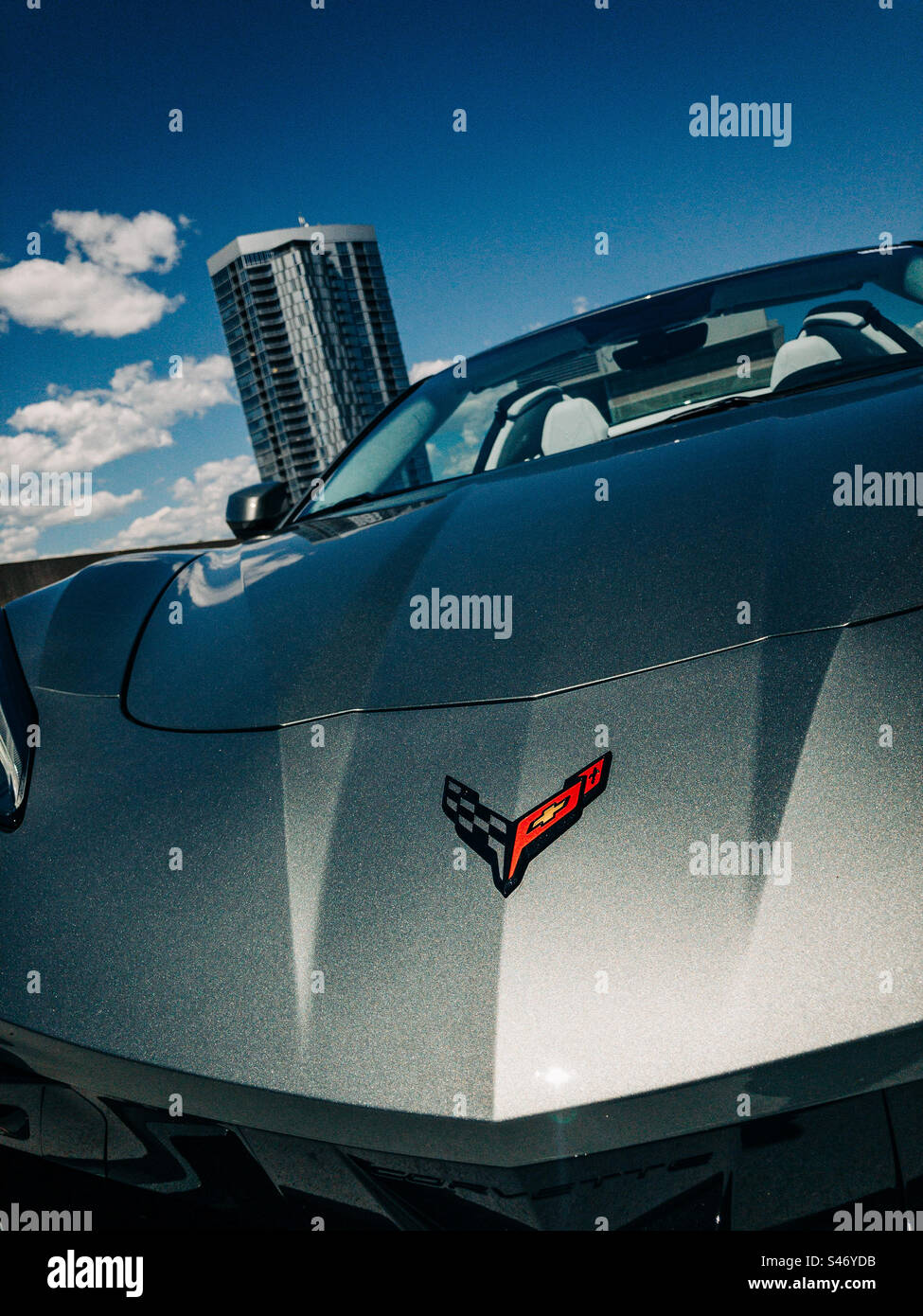 Close-up photo of the logo on a silver 2023 Chevrolet Corvette Stingray, 70th Anniversary Edition, sitting on the top floor of a parking garage in Denver, CO, with buildings, sky, and clouds. Stock Photo
