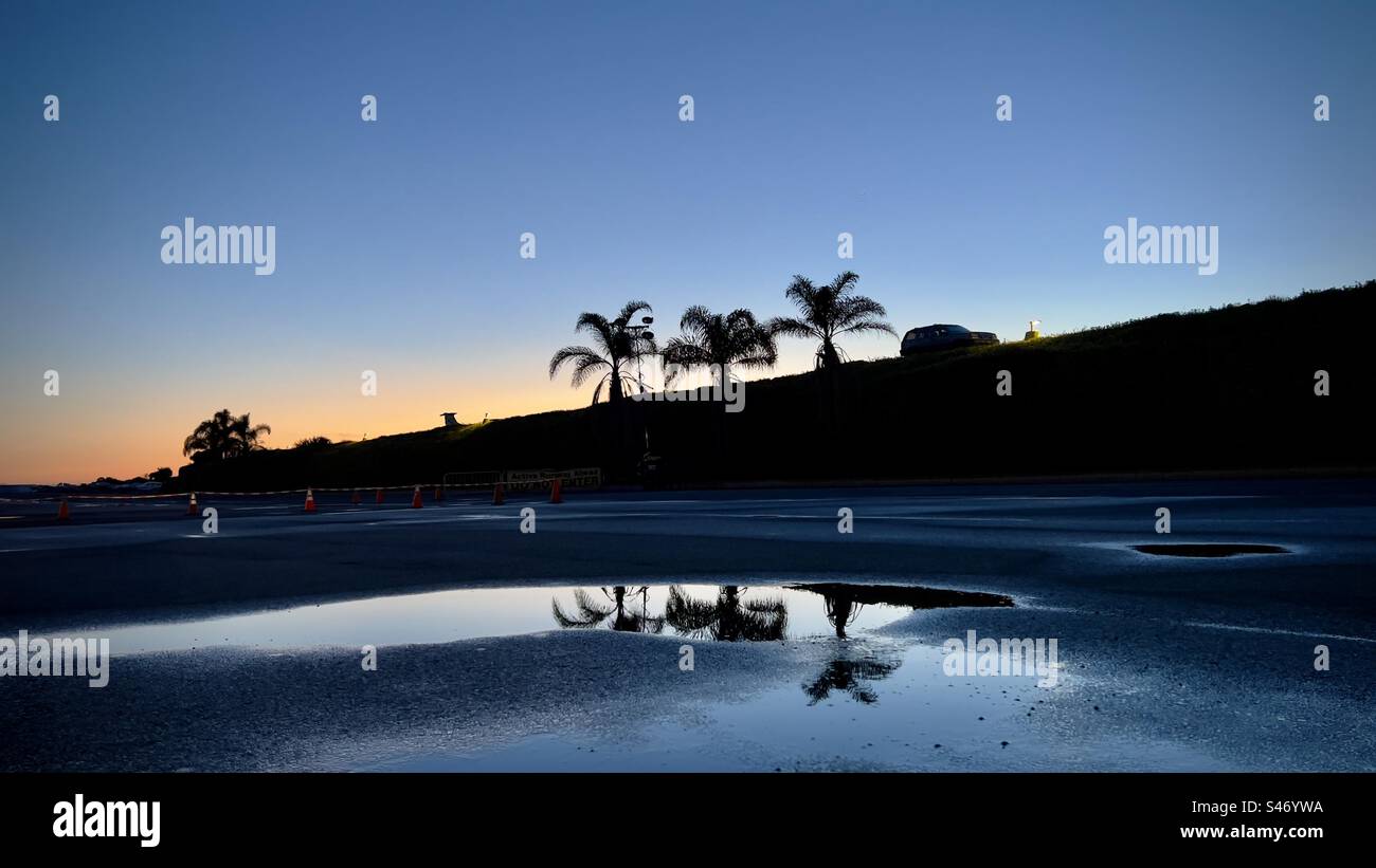Wide view, silhouetted SUV and palm trees at top of hill with orange sunset sky in background and reflections on puddles in parking lot in foreground Stock Photo