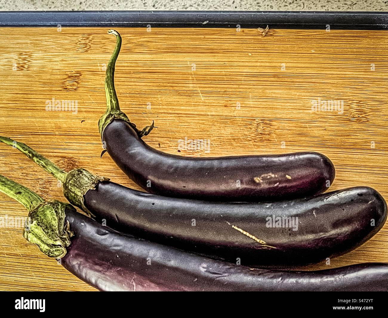 High angle, close-up view of three Lebanese eggplants on wooden cutting board on kitchen counter with copy space. Healthy eating. - Smartphone Captured Stock Image
