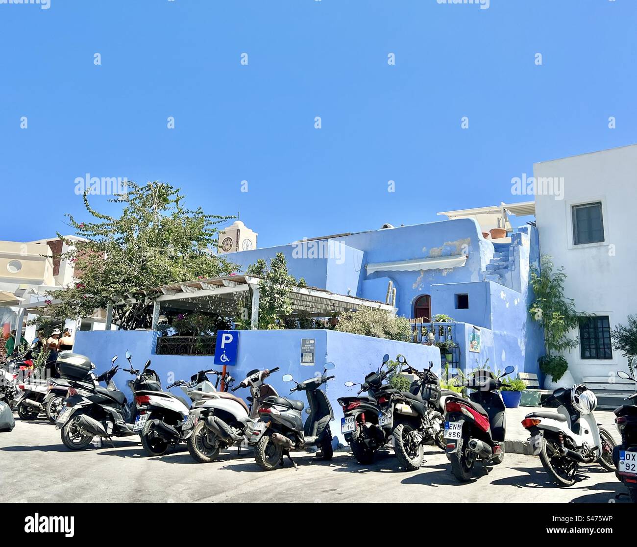 Motorcycles and mopeds in a parking area located next to a vibrant blue building that matches the sky on a summer day, Oia, Santorini. - Smartphone Captured Stock Image
