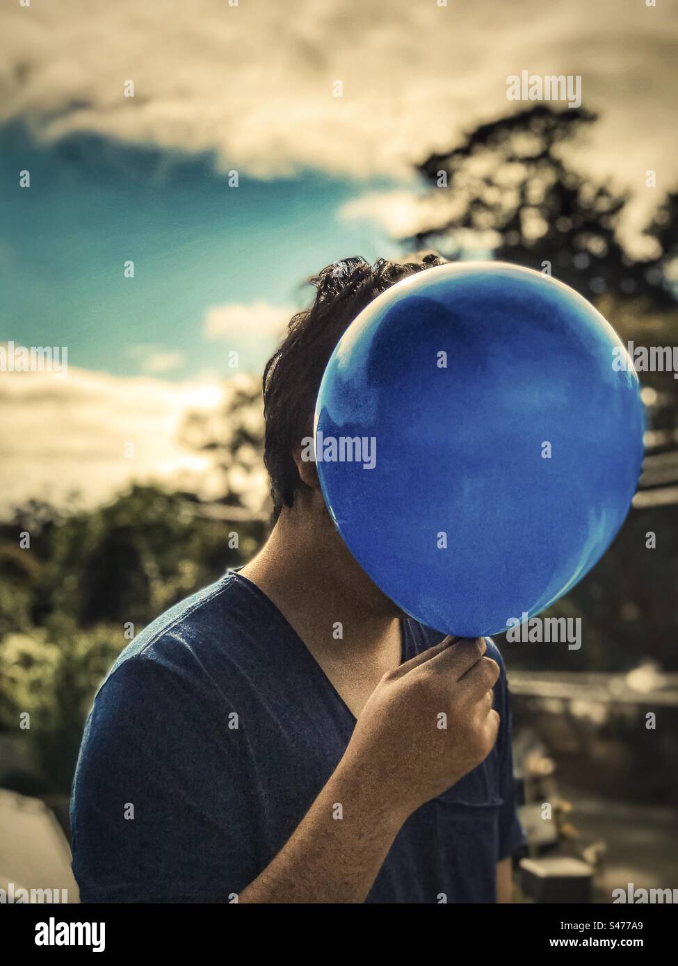 Blue mood day. Conceptual shot of young man holding blue balloon against moody background of blue sky, white clouds and trees. Obscured face. Depression. Mental health. - Smartphone Captured Stock Image
