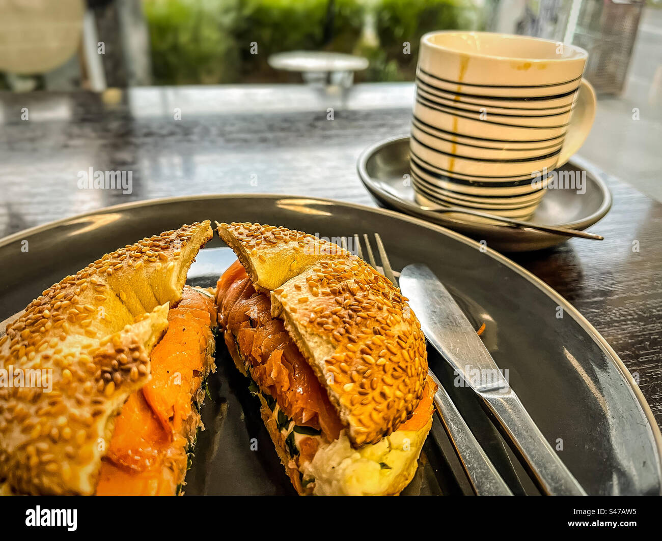 Close-up of salmon and cream cheese bagel on plate with cutlery and coffee cup on table in front of window at cafe. - Smartphone Captured Stock Image