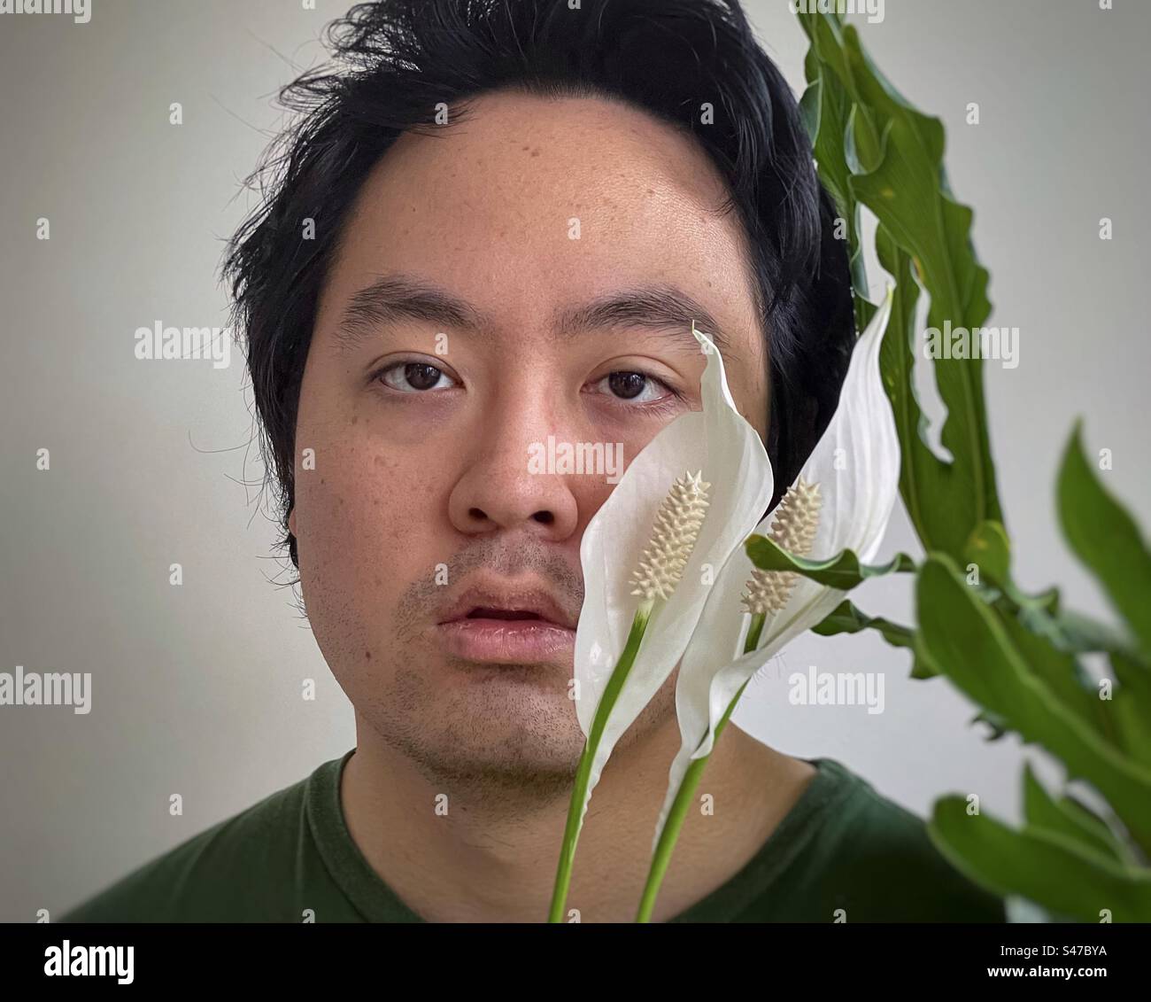 Close-up headshot of young Asian man framed by two peace lily flowers and philodendron leaves. - Smartphone Captured Stock Image