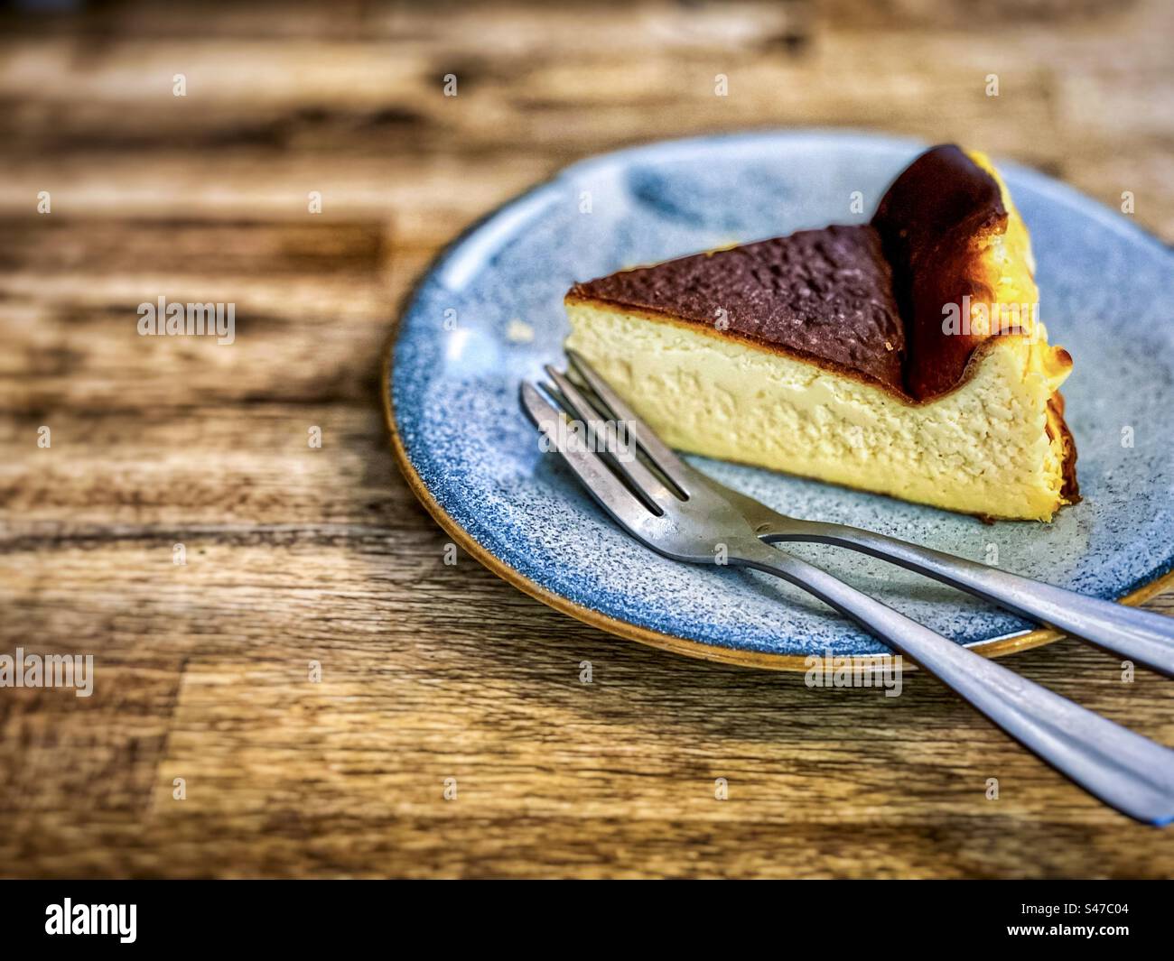 Close-up of a slice of Basque cheesecake on a plate with two forks on wooden table. - Smartphone Captured Stock Image