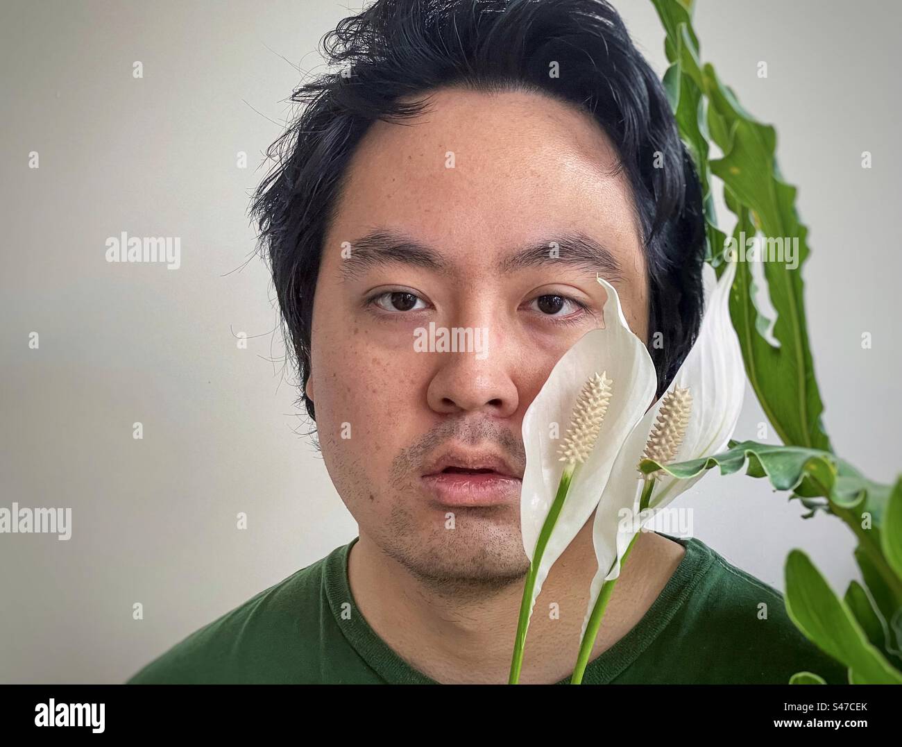 Close-up portrait of young, Asian man framed by two peace lily flowers and philodendron leaves with copy space. - Smartphone Captured Stock Image