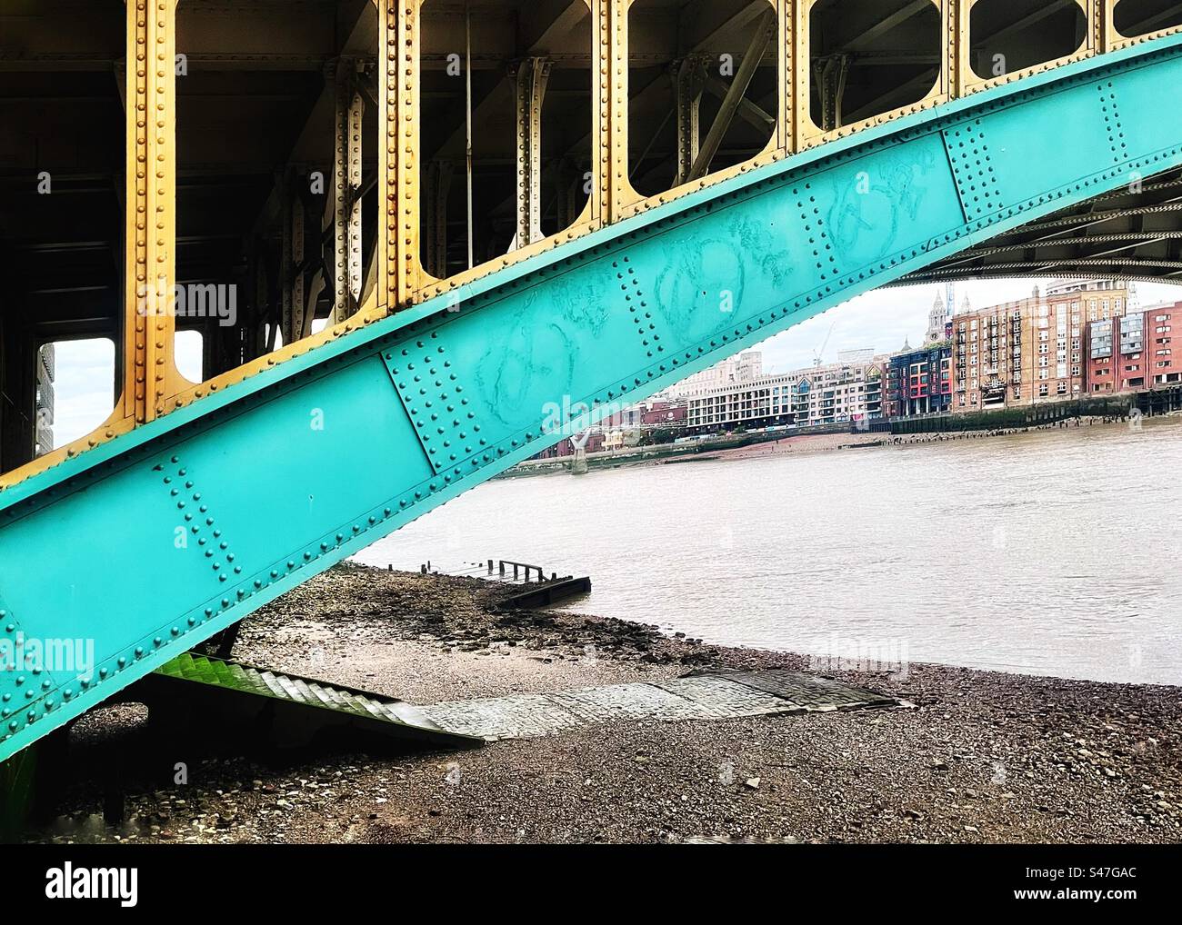 Underneath the arch of Southwark Bridge in London with the River Thames at low tide exposing the shingle and shoreline. Launching steps for boats pictured. - Smartphone Captured Stock Image