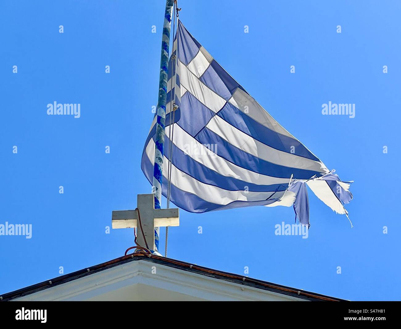 The blue and white flag of Greece behind a white cross top a church roof, surrounded by blue summer sky. Megalochori, Santorini Stock Photo