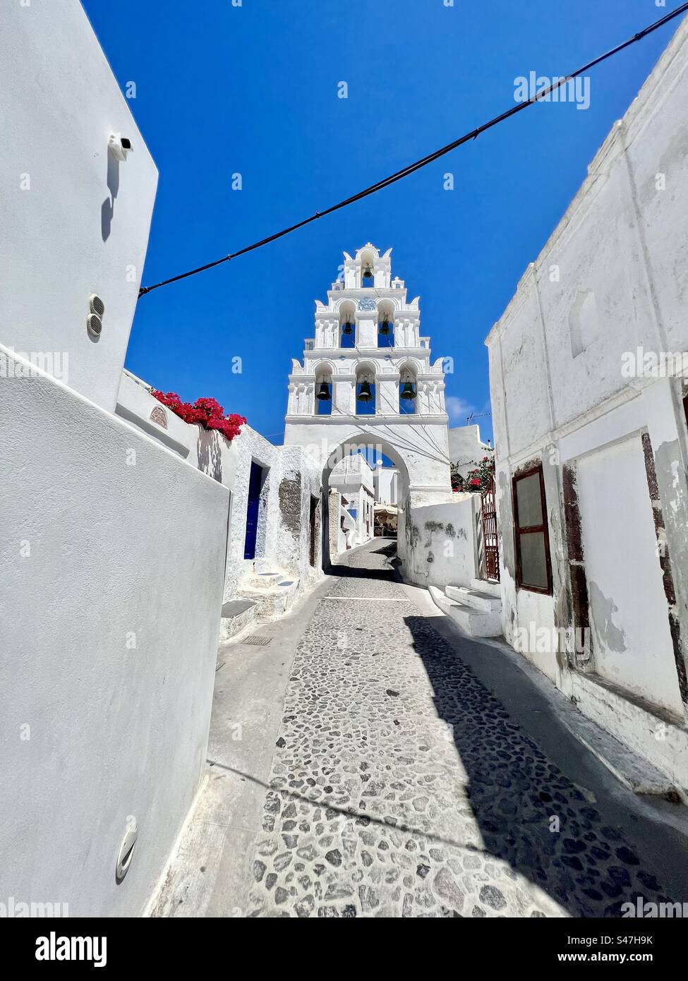 Bell tower of Megalochori, with 6 bells and arched opening, over a narrow cobblestone street near the center of this traditional village on Santorini. - Smartphone Captured Stock Image
