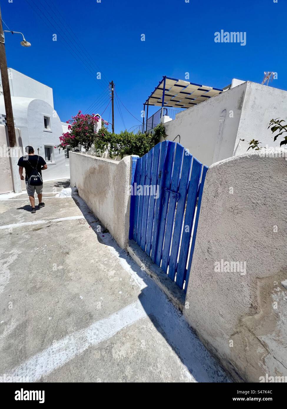 A blue painted gate and young man walking down a narrow street in Akrotiri village of Santorini island. - Smartphone Captured Stock Image