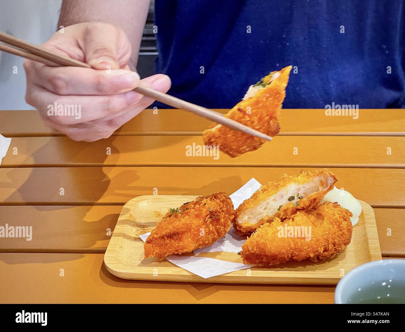 Midsection of man using chopsticks to pick up a piece of Japanese shrimp croquette from plate on wooden table. Japanese cuisine, korokke. - Smartphone Captured Stock Image