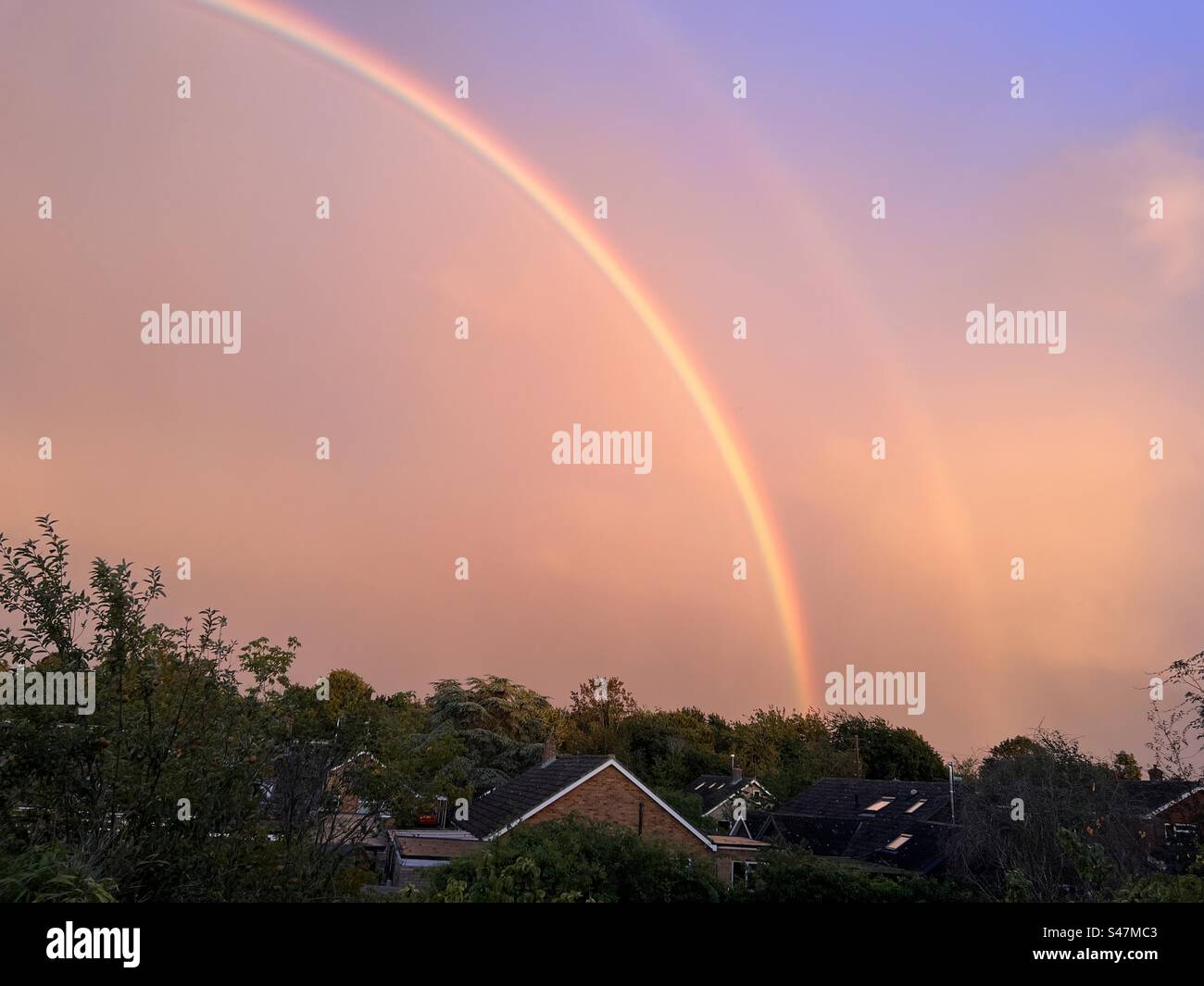 Sunset landscape with beautiful rainbow across sky turning it pink and orange over roof tops and fields in Summer - Smartphone Captured Stock Image