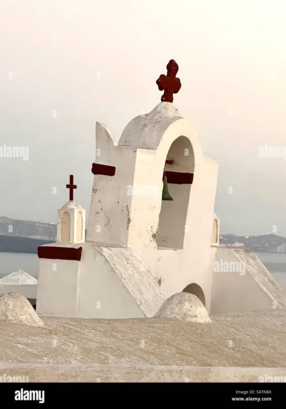 A whitewashed bell tower with brown accents and a cross sit atop a building of the caldera in Oia against a dusk sky on a summer eve. - Smartphone Captured Stock Image