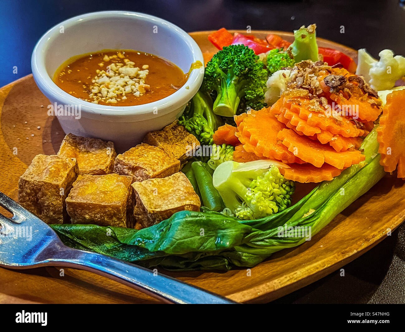 Close-up of crispy fried tofu cubes and freshly steamed vegetables with a spicy peanut sauce dip in bowl on plate on table. Vegetarian/ vegan food. Healthy eating. Eat your colors. - Smartphone Captured Stock Image