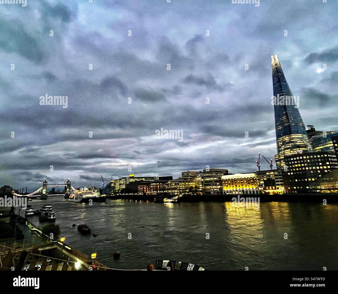 Stormy skies in London, looking at The Shard and Tower Bridge from London Bridge across the River Thames at dusk with lights on - Smartphone Captured Stock Image