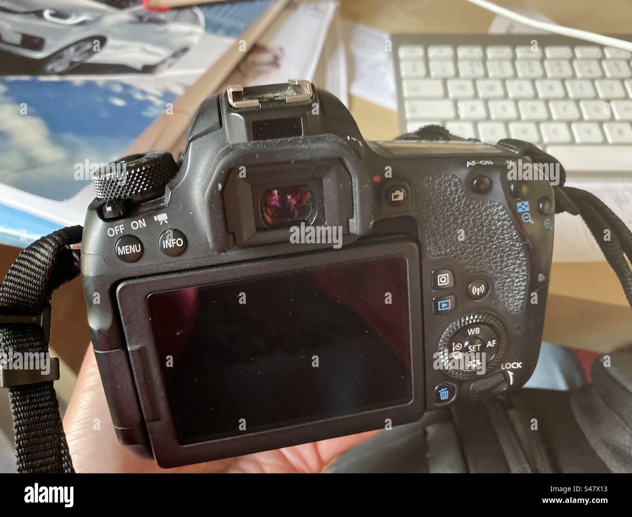 Hand holding a DSLR camera above a cluttered work desk with papers and keyboard - Smartphone Captured Stock Image