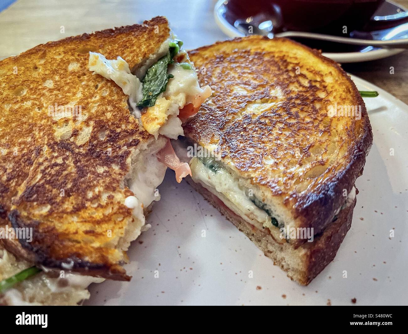 Close-up of partly eaten haloumi, spinach and tomato toastie on plate on table with a coffee cup and saucer. Comfort food. Breakfast. - Smartphone Captured Stock Image