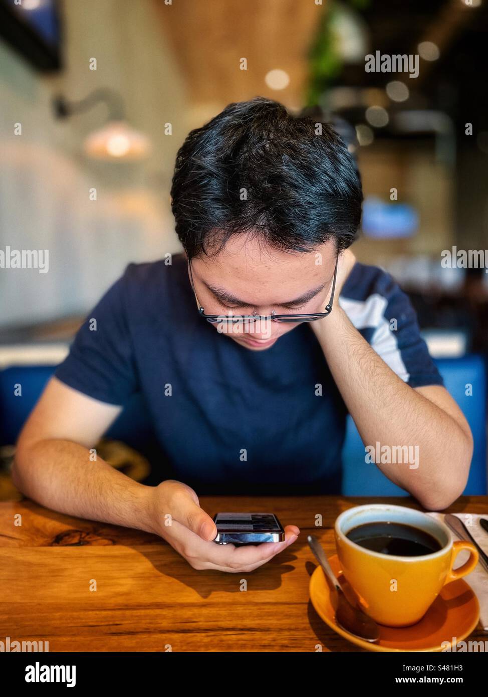 Young Asian man in eyeglasses using mobile phone while drinking a cup of black coffee at table in a cafe. Food and drink. Wireless technology. Focus on foreground. - Smartphone Captured Stock Image