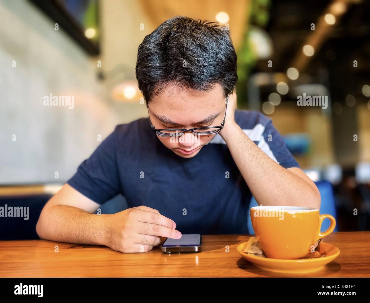 Young Asian man in eyeglasses using a mobile phone while drinking coffee at a table in a cafe. Wireless technology. Lifestyle. Food and drink. Focus on foreground. - Smartphone Captured Stock Image