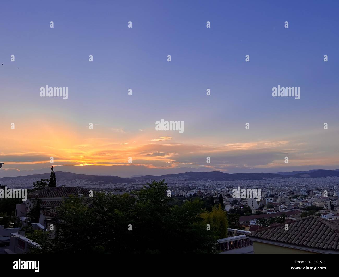 The rays of the sun spread across the blue evening sky as it sinks into the Athens horizon at the end of the daytime. - Smartphone Captured Stock Image
