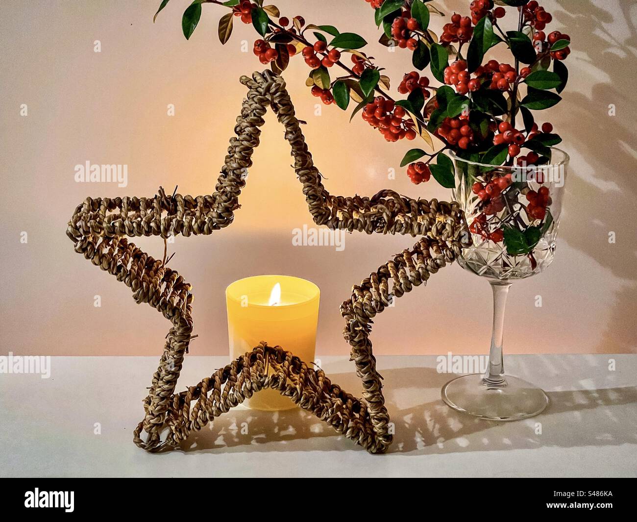 Arrangement of lighted candle behind decorative rattan star and red Cotoneaster berry branches in crystal goblet. Illuminated. Christmas. Celebration. - Smartphone Captured Stock Image