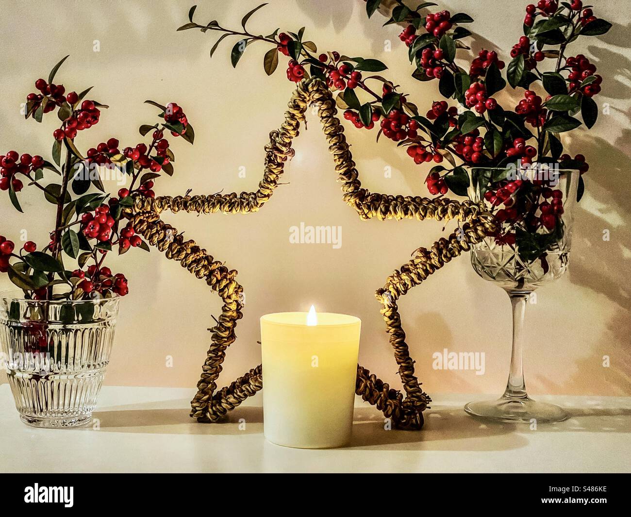 Lighted candle in front of decorative Christmas rattan star with red Cotoneaster berry branches in crystal glasses with illumination. Christmas. Celebration. Decoration. Still life. - Smartphone Captured Stock Image