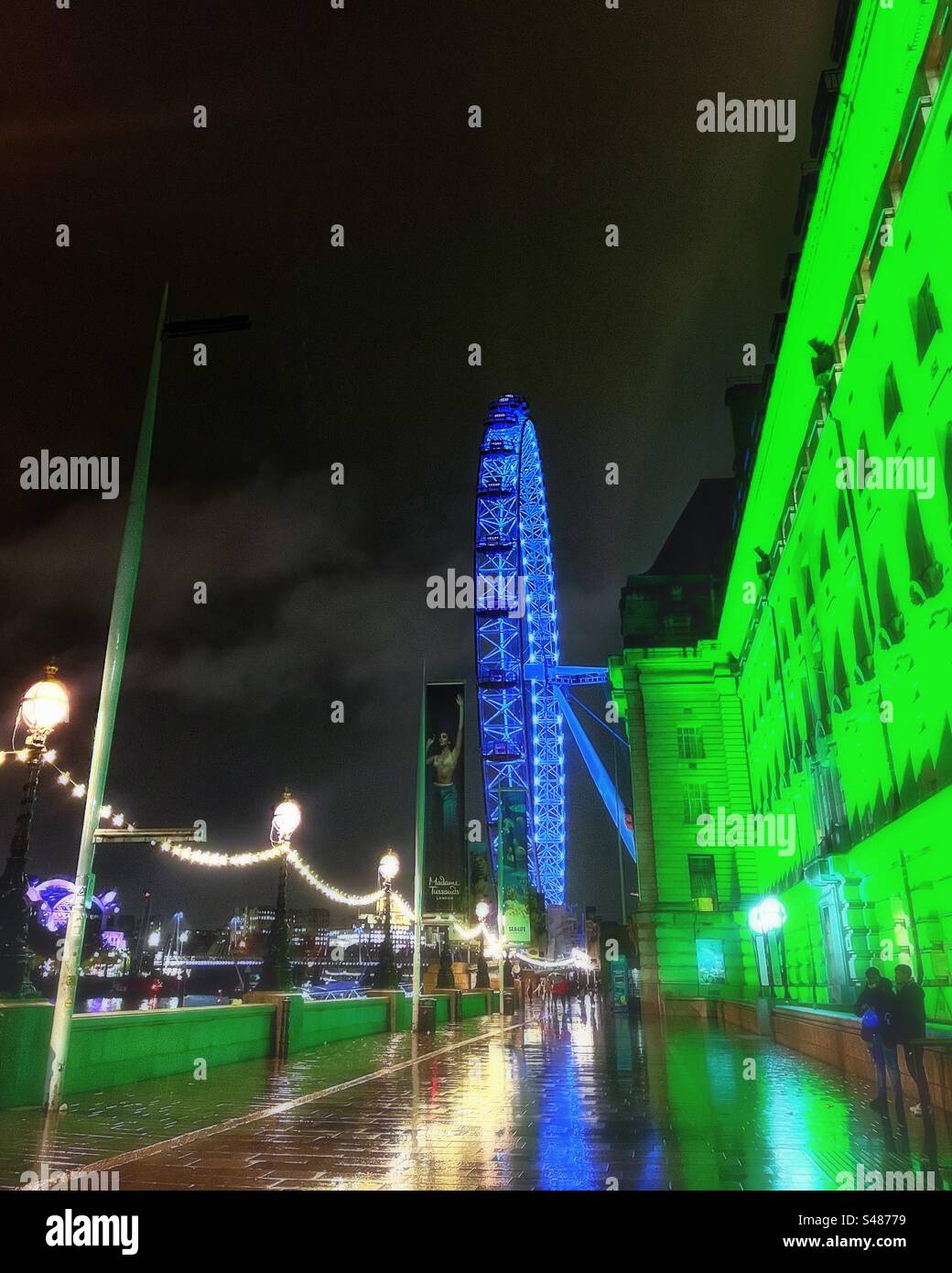 A view from Westminster Bridge towards the London Millennium Wheel aka London Eye including County Hall and heritage lights illuminated at night with reflections in the rain - Smartphone Captured Stock Image