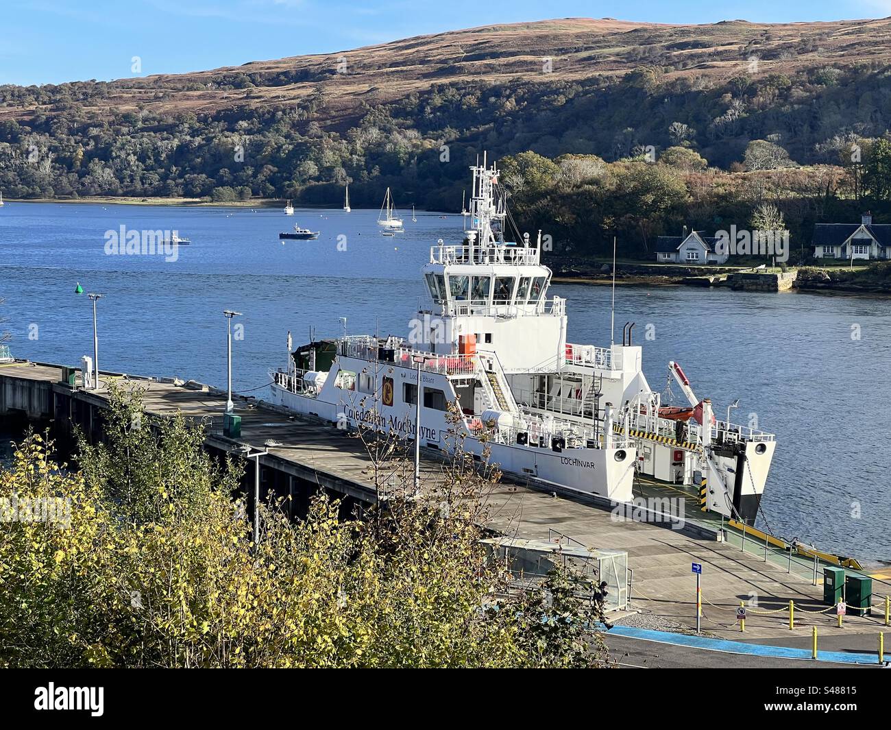A ferry moored in Loch Aline harbour on a calm autumn day, with the loch and sailing boats visible in the background. The ferry runs between Loch Aline and Fishnish. Stock Photo