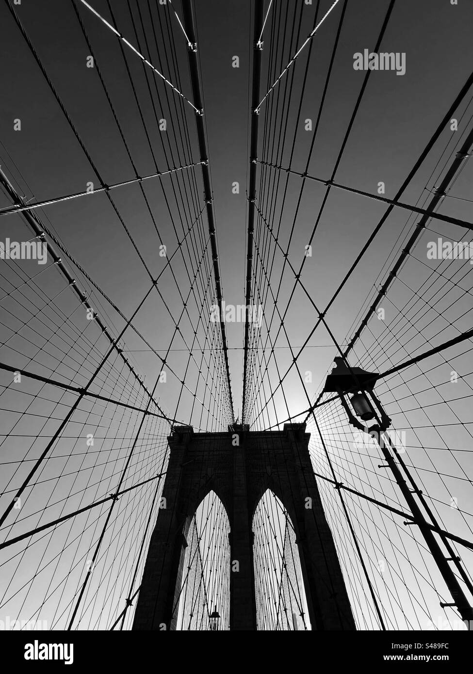 Arch support of Brooklyn Bridge in silhouette with pattern of suspension cables and lamp. Backlit by the sun behind the bridge tower support - Smartphone Captured Stock Image