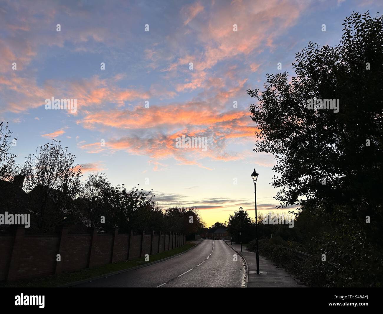 Sunset over suburban UK landscape. Blue sky with salmon pink and orange clouds. Silhouetted trees and lampposts. - Smartphone Captured Stock Image