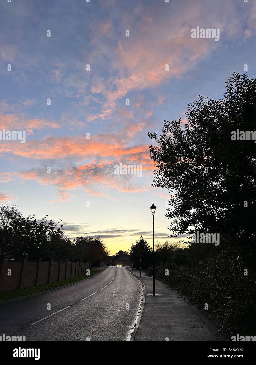 Sunset over suburban UK landscape. Blue sky with salmon pink and orange clouds. Silhouetted trees and lampposts. - Smartphone Captured Stock Image