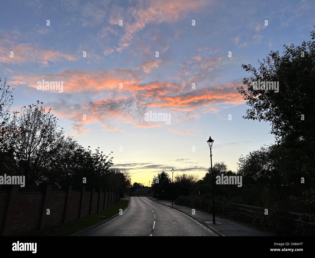 Sunset over suburban UK landscape. Blue sky with salmon pink and orange clouds. Silhouetted trees and lampposts. - Smartphone Captured Stock Image