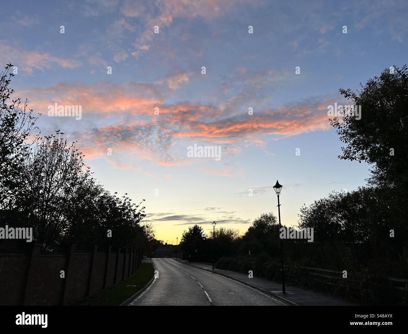 Sunset over suburban UK landscape. Blue sky with salmon pink and orange clouds. Silhouetted trees and lampposts. - Smartphone Captured Stock Image