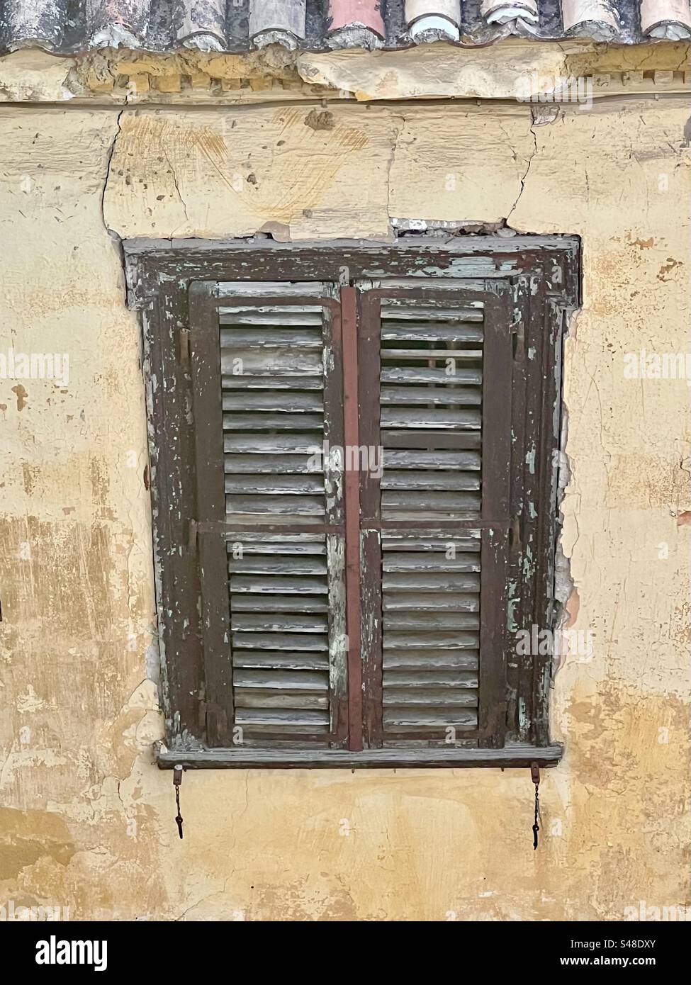 Painted shutters with peeling and chipping brown paint closed on a window of tan building - Smartphone Captured Stock Image