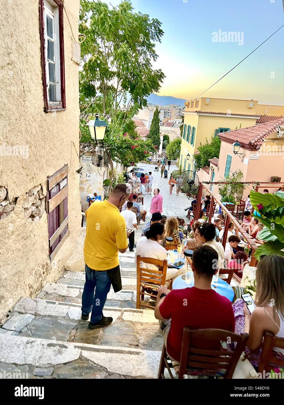 Colorful scene at sunset in Athens on a small street lined with tables and chairs of a cafe that fits amongst the steep steps of the incline. - Smartphone Captured Stock Image