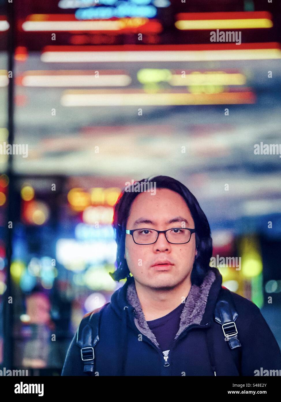 Young Asian man in eyeglasses standing in front of glass shop window with reflections of neon lights in the city at night. - Smartphone Captured Stock Image