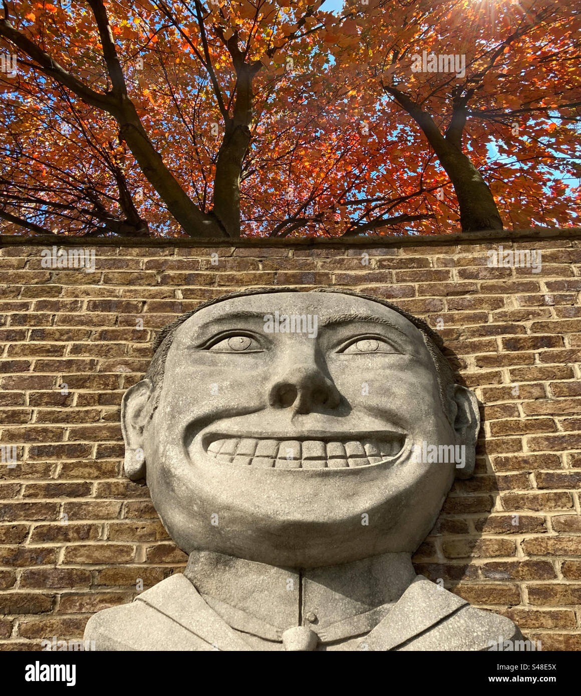 Relief sculpture of a grinning face on a brick wall with autumn trees branching overhead Stock Photo