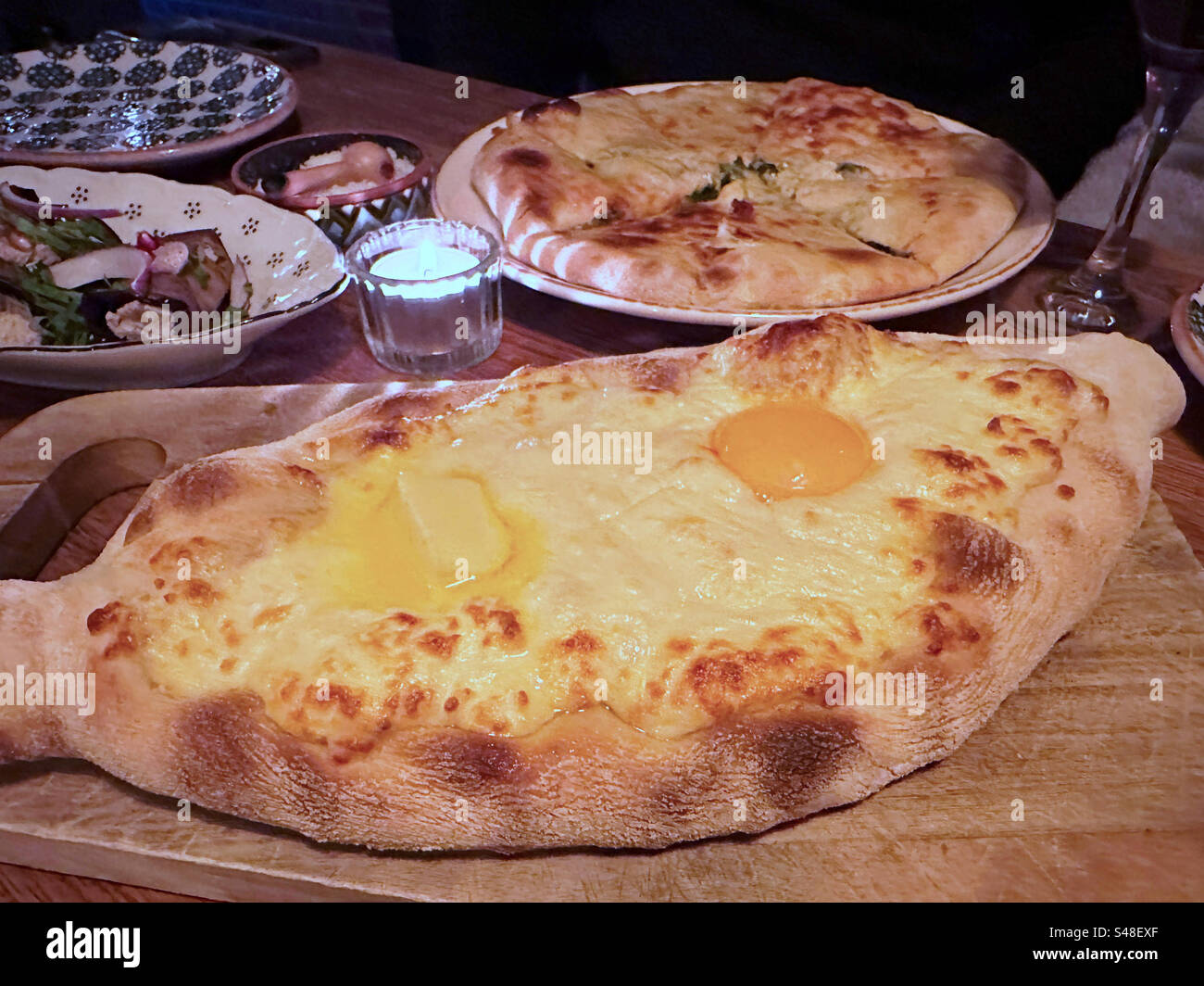 Khachapuri (Georgian Cheese Bread)served with a raw egg and butter which are then mixed into the hot cheese to cook.  In the background is a spinach pie. - Smartphone Captured Stock Image