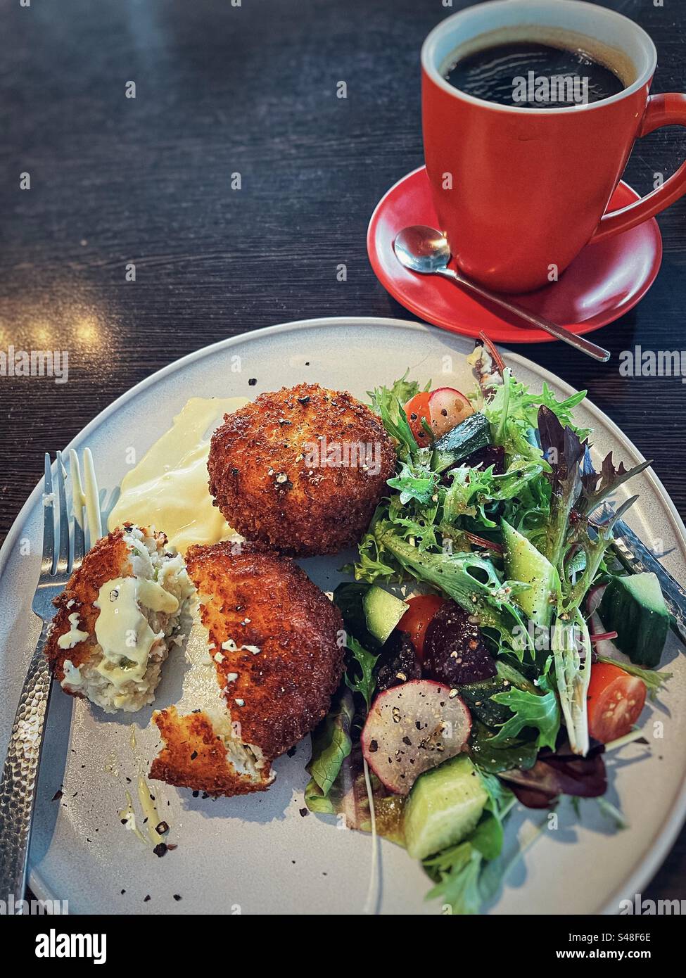 High angle view of salmon patties with hollandaise sauce and fresh salad on plate with cutlery and black coffee in red mug and saucer on table. - Smartphone Captured Stock Image