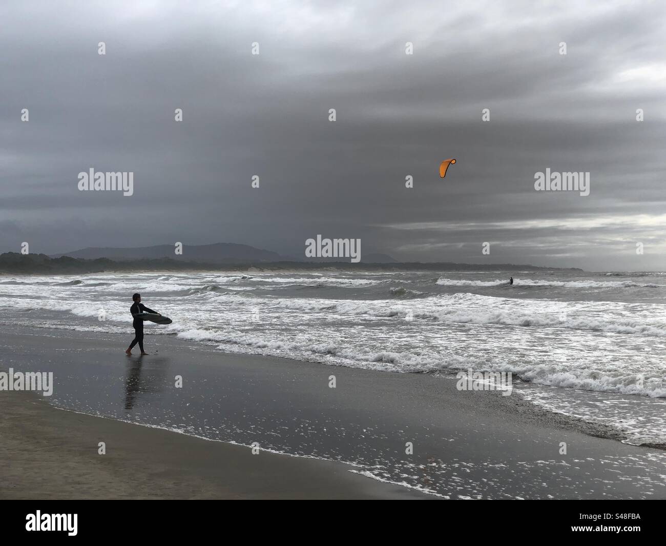 Cabedelo, Portugal - November 11, 2023: A surfer entering the water of Atlantic Ocean as another surfer riding an orange kite - Smartphone Captured Stock Image
