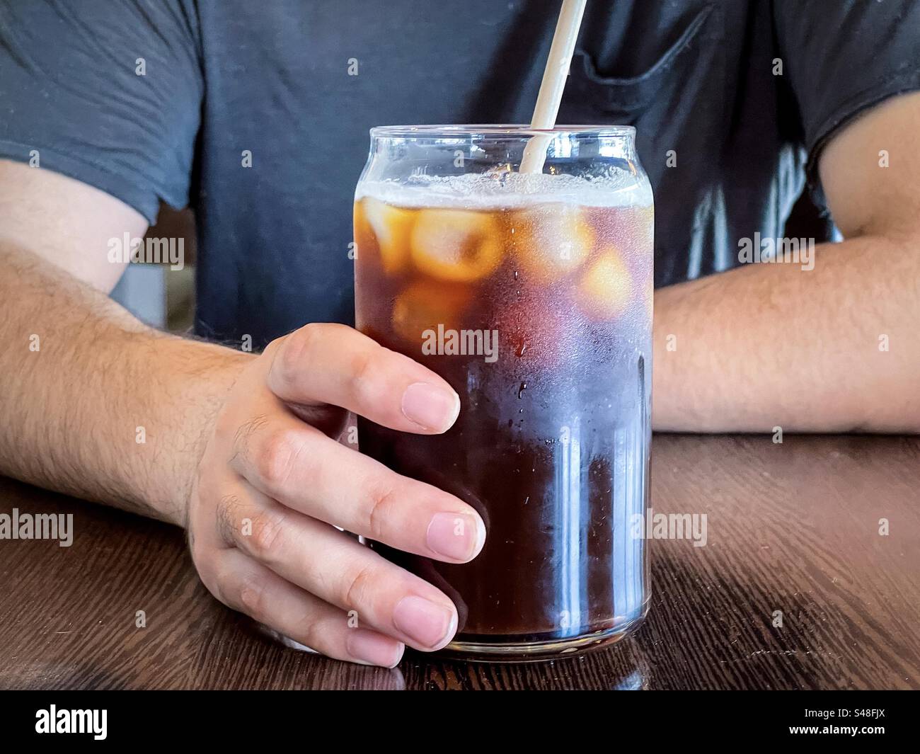 Midsection of man with one hand holding a glass of iced coffee with a straw on table. - Smartphone Captured Stock Image