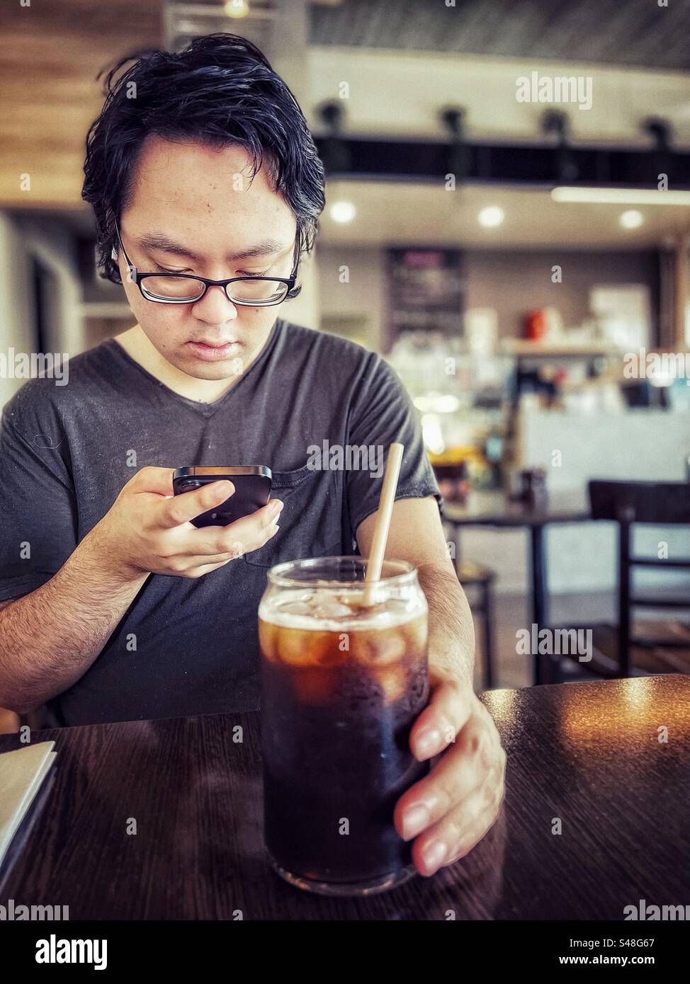Asian man in eyeglasses using a mobile phone while drinking iced coffee at table in a cafe. Wireless technology. Communication. Food and drink. - Smartphone Captured Stock Image
