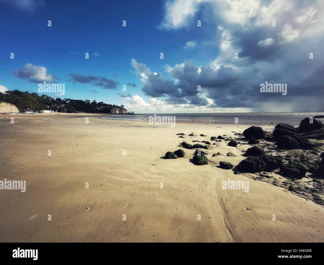 Wide empty sandy beach with black small rocks and headland with a blue sky and storm clouds at Meschers sur Gironde in the Charente Maritime Stock Photo