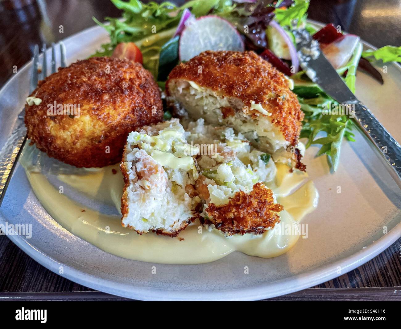 Close-up of salmon patties with hollandaise sauce and fresh salad on plate with cutlery on table. - Smartphone Captured Stock Image