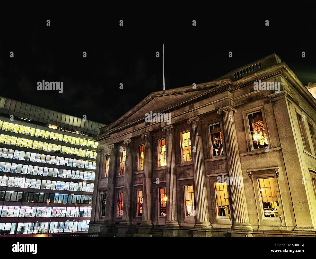 View of the Fishmongers' Hall next to London Bridge at night, windows illuminated and chandeliers visible. Fish Hall is a Listed building; adjacent office block contrasting architecture - Smartphone Captured Stock Image
