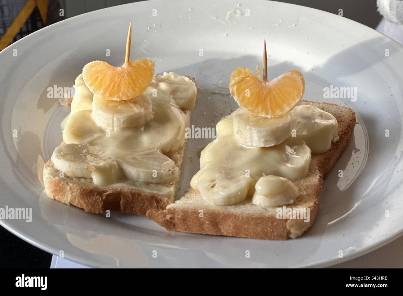 Two banana cream sandwiches on a white plate. Orange or clementine slices are stacked on a toothpick atop bananas and vanilla cream on toasted bread. - Smartphone Captured Stock Image