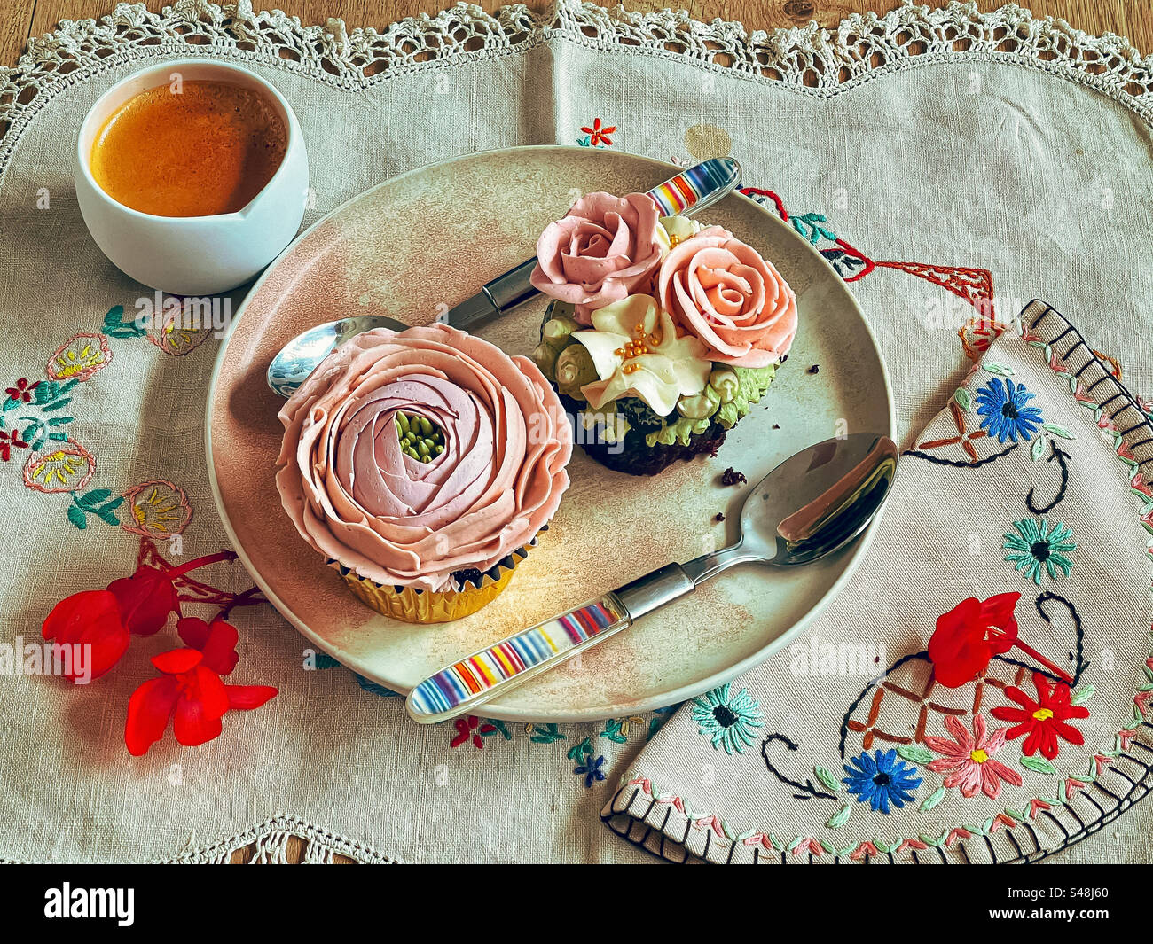 High angle view of two cupcakes with buttercream frosting flowers on plate with spoons and a cup of expresso coffee on vintage, embroidered tablecloth. Vintage style teatime. - Smartphone Captured Stock Image