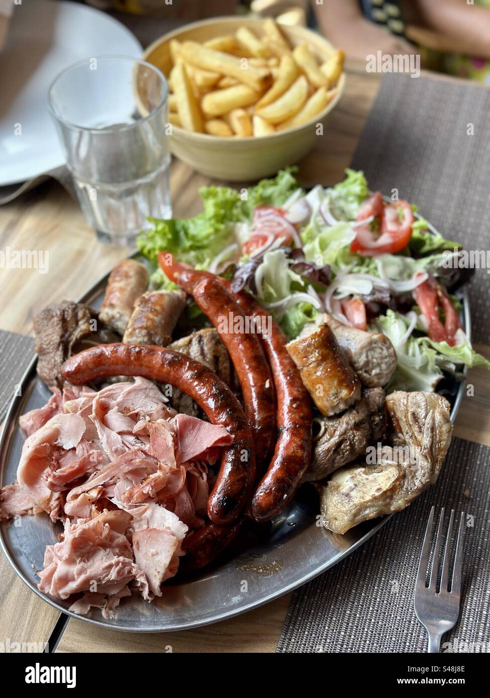 Close-up shot of a huge mixed grill platter on a silver plate on a restaurant table, with sausages, salad, steak, fries and a fork. - Smartphone Captured Stock Image