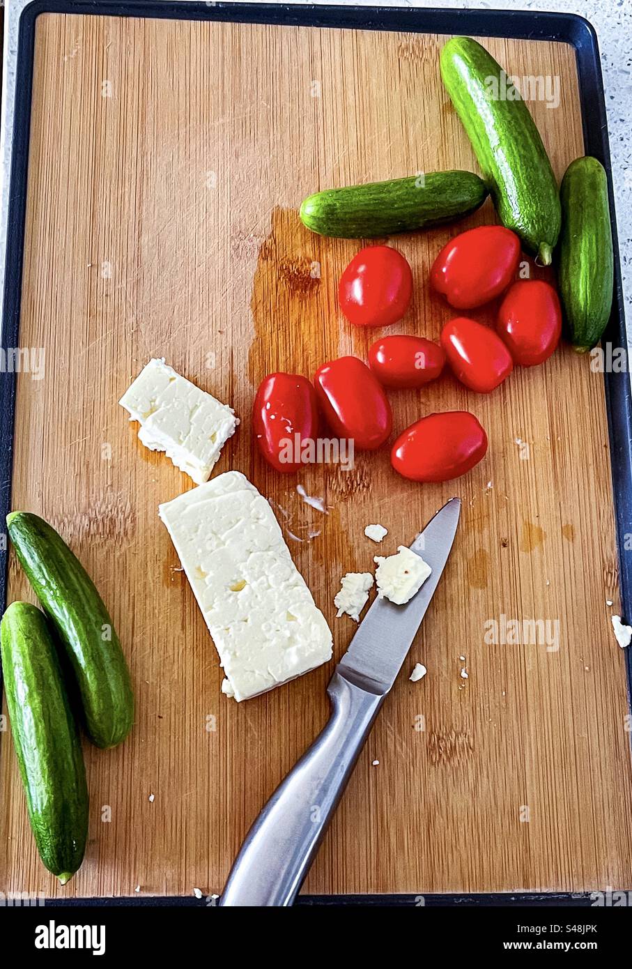 High angle view of sliced feta cheese, cherry tomatoes, baby cucumbers and knife on wooden cutting board on kitchen counter. Healthy eating. Raw food diet. Vegetarian food. Food preparation. - Smartphone Captured Stock Image