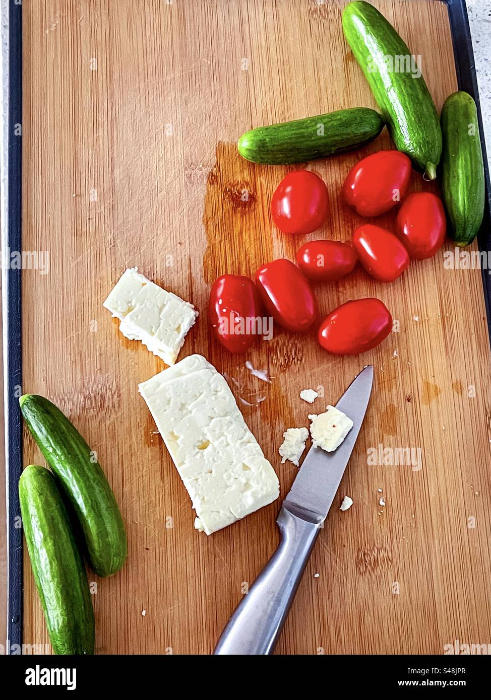 High angle view of sliced feta cheese, cherry tomatoes, baby cucumbers and knife on wooden cutting board on kitchen counter. Healthy eating. Raw food diet. Vegetarian food. Food preparation. - Smartphone Captured Stock Image